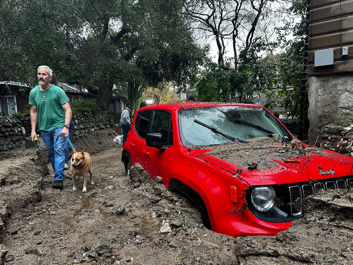 A resident and their dog walk past a vehicle partially submerged in mud after a storm Friday, Feb. 14, 2025, in Sierra Madre, Calif.