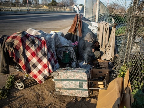 A man starts a fire while his wife sleeps inside their makeshift tent along a barbed wire fence near Highway 99 in southwest Fresno on Feb. 11, 2022.