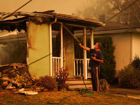 A person uses a hose to spray water on the flames of a house to prevent the Eaton Fire from spreading to other homes in Pasadena on Jan. 8, 2025. State Farm says losses from the fire have weakened its financial position.