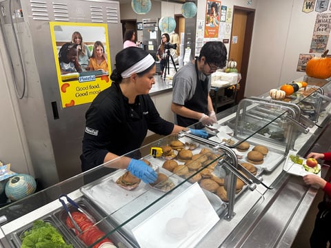 Kitchen staff at Lincoln Crossing Elementary serve up a burger lunch with sides of fruits and veggies on October 27, 2025. Since partnering with the nonprofit Eat Fresh, over half of the school meals are now made up of freshly prepared food.