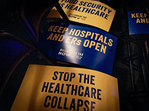 Signs reading “A Billionaire Tax Now” rest on chairs at a Billionaire Tax Now rally where Sen. Bernie Sanders is set to speak in Los Angeles on Feb. 18, 2026.