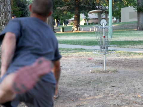 A player throws a disc toward a metal basket at the Oak Park disc golf course.