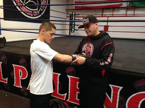 On the left, Brent Venegas III slides his hand into a ten-ounce boxing glove held by trainer Gabriel Flores Sr.