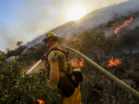 A firefighter douses the grass with water along a hillside on a wildfire in Azusa, Calif., Monday, June 20, 2016. Police in the city of Azusa and parts of Duarte ordered hundreds of homes evacuated. Others were under voluntary evacuations.