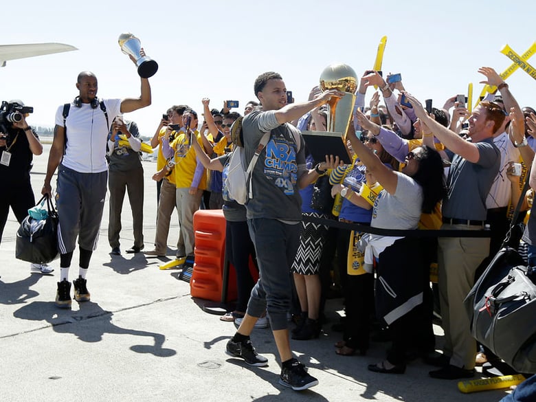Golden State Warriors guard Stephen Curry, center, walks past cheering team employees as he carries the Larry O'Brien championship trophy in front of forward Andre Iguodala, lifting the NBA Finals MVP trophy, after the team landed in Oakland, Calif.